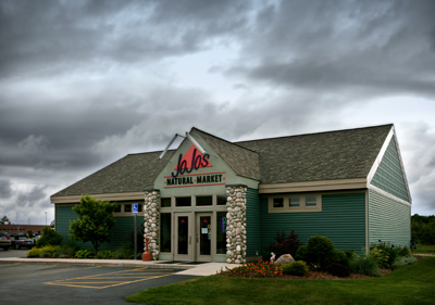 picture of Jo Jo's Natural Market - specialty food store in Gaylord, Michigan - dark green wood siding, grey-green architectural shingled roof, white trim, fieldstone columns at entrance