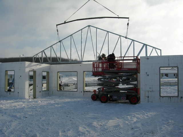 picture of steel roof truss being set in place on ICF walls, with workman on scissor-lift guiding installation and ensuring a proper fit