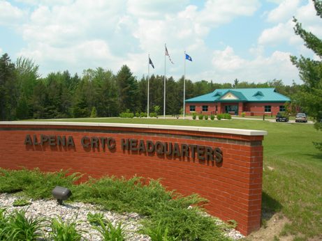 exterior picture of Headquarters Building at Alpena CRTC with flags - photo taken from the entrance road in front of the building's curved masonry wall sign - the building has red masonry walls, green metal roof and trim