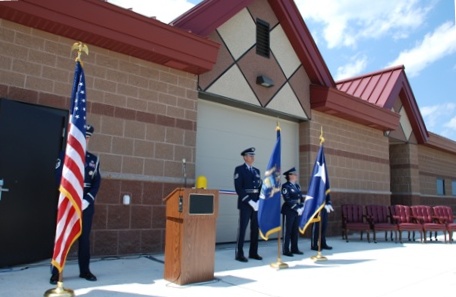 this is a photo taken the day of the Ribbon Cutting Ceremony (Summer 2008) which officially opened the facility for operation - Armed Forces personnel in dress uniforms standing at parade rest with flags flanking speaker's podium - building has concrete masonry unit walls in multiple tones of tan and brown with red metal roof