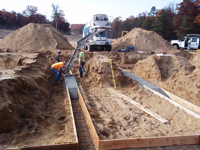 Concrete truck in background pours concrete into footing forms at excavated site
