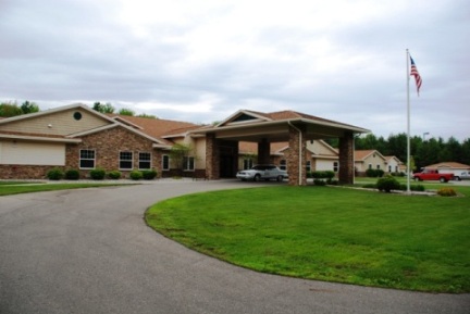 exterior photo of Tawas Village Assisted Living Facility - curved entrance drive through porte-cochere, the buildings are brick with tan siding, white trim and earth-tone shingled roofs