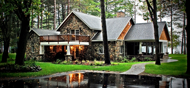 exterior photo of a fieldstone home with cedar shakes, white window and soffit trim, and a walk out deck over the covered side entry.  The picture was taken in early Autumn, while a light rain was falling, turning the driveway into a dark mirror reflecting the porch lights.