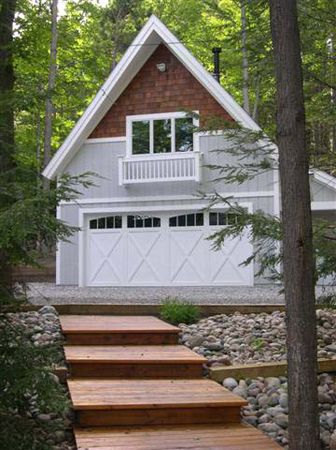 picture of detached garage for a waterfront home on Kassuba Lake  grey siding, white trim, cedar shakes at second floor roof peak, custom garage doors - wood walkway weaves through trees and beds of river rock to garage