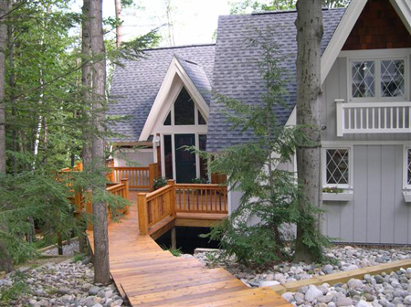 picture of a customized 'A' frame styled cottage with grey siding and roof, white trim and natural wood multi-level decks and walkways - the home appears to be springing from beds of river rock, nestled in a mixed-stand of mature trees
