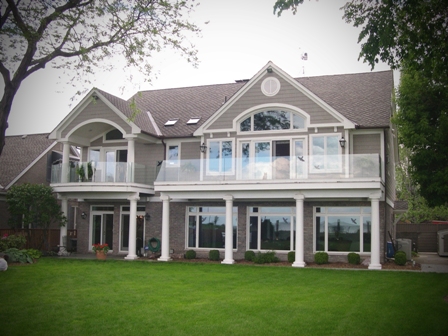 exterior photo of the lakeside elevation of custom waterfront home on Lake St. Clair, MI showing upper level balconies with glass wall safety railing - upper balconies create a covered porch area for the lower level of the home