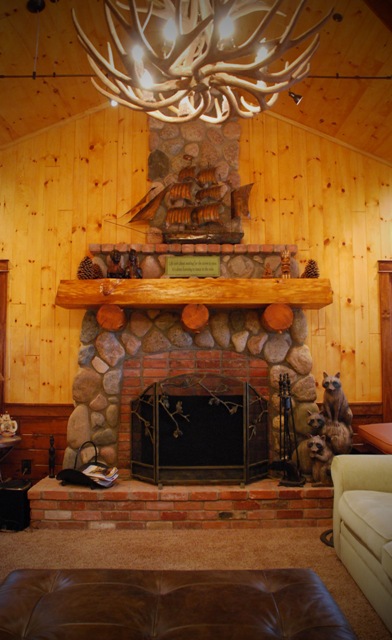 interior photo of living room with fieldstone fireplace, timber mantle, wood plank walls, ceiling and antler pendant light