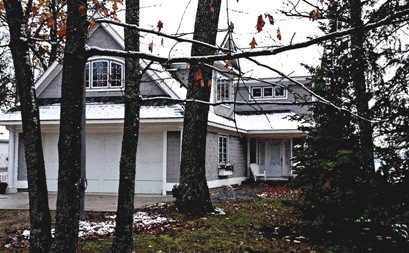 picture of lakeside home in Topinabee, Michigan - this exterior photo is looking through trees at the home's main entry showing snow-dusted roof, dark grey cedar siding and white trim, white wood-and-glass entry door and sidelights, and custom windows for bonus room above attached garage