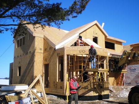 picture of workers installing exterior framing on a custom home being built on Otsego Lake in Gaylord Michigan - the snow covered lake is visible in the background