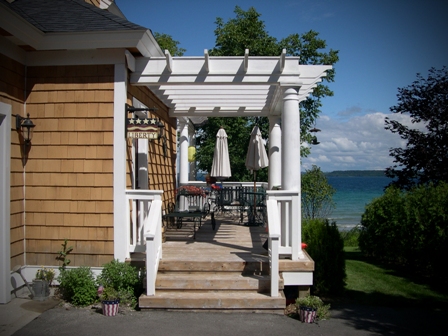 picture of custom lakefront cottage on West Bay, Traverse City, MI - this exterior photo is of the side entrance and deck of the cottage with natural cedar siding, white trim and pergola construction on porch, with view of Traverse Bay beyond
