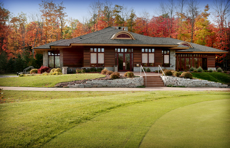 exterior picture of UAW Black Lake Golf Course Clubhouse - dark wood siding, wood trim accents, grey shingle roof - picture taken from on the green looking toward clubhouse with backdrop of trees with Autumn foliage