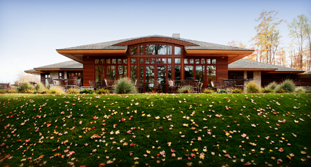 exterior picture of UAW Black Lake Golf Course Clubhouse - looking at window wall facing greens covered with autumn leaves, building has dark wood siding, stone and wood accents and trim, with grey shingle roof